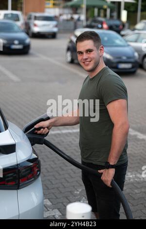 Ein lächelnder Mann lädt ein Elektroauto an einer Ladestation auf einem öffentlichen Parkplatz auf. Stockfoto