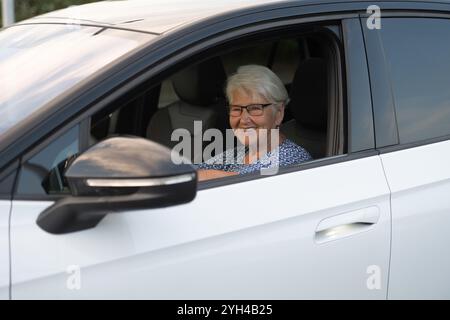 Eine fröhliche ältere Frau mit Brille sitzt auf dem Fahrersitz eines weißen Autos und blickt aus dem offenen Fenster. Stockfoto
