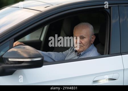 Ein nachdenklicher älterer Mann in einem karierten Hemd sitzt auf dem Fahrersitz eines weißen Autos und blickt ruhig aus dem Fenster. Stockfoto