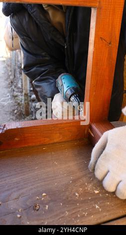 Vertikales Foto des Zimmermanns, der Löcher in braunem Holzrahmen bohrt und Löcher für Befestigungselemente in Holzleisten mit Bohrer herstellt Stockfoto