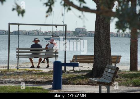 Blick von hinten älteres Paar mit Hüten auf einer Metallschaukel über den Sandstrand hinaus zu Segelbooten am Gulfport Florida Beach. Gebäude und ja Stockfoto