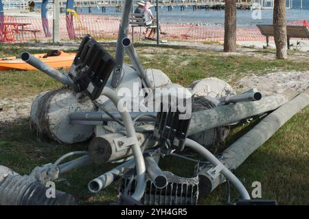 Ein Haufen beschädigter Metall- und Betonstangen im Vordergrund hinter einem älteren Paar, das Hüte trägt und auf einer Metallschaukel über dem Sandstrand sitzt und zum Segeln hinausgeht Stockfoto