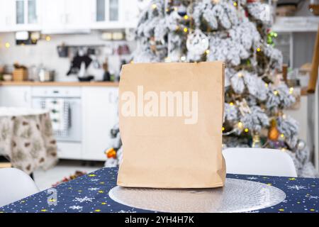 Auf dem Küchentisch liegt eine Kraftpapiertüte. Lieferung von Sushi und Brötchen, Keksen, Geschenk im festlichen Innenraum des Hauses mit einem weißen Weihnachtsbaum Stockfoto