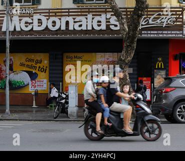 Hanoi, Vietnam - 5. Juli 2023: Familienfahrt mit dem Motorrad vorbei an einem McDonald's Fast Food Restaurant in einer geschäftigen Straße in der Innenstadt von Hanoi. Stockfoto