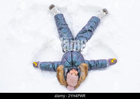 Ein Mädchen in einem blauen Jumpsuit macht Schneeengel im Schnee. Winterspaß, Spiele im Freien, Weihnachten Stockfoto