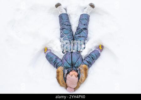 Ein Mädchen in einem blauen Jumpsuit macht Schneeengel im Schnee. Winterspaß, Spiele im Freien, Weihnachten Stockfoto