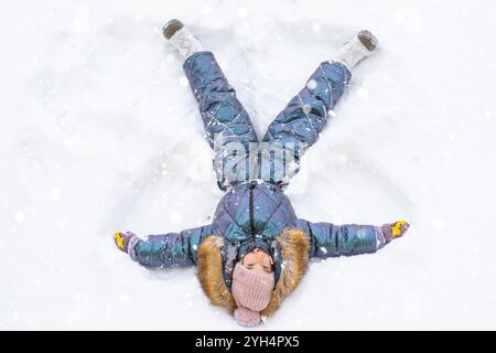 Ein Mädchen in einem blauen Jumpsuit macht Schneeengel im Schnee. Winterspaß, Spiele im Freien, Weihnachten Stockfoto