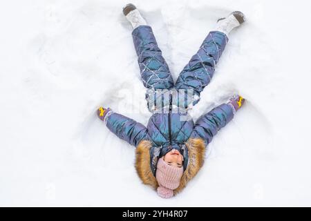 Ein Mädchen in einem blauen Jumpsuit macht Schneeengel im Schnee. Winterspaß, Spiele im Freien, Weihnachten Stockfoto