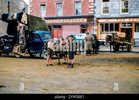 Der Fair Day in Castleisland im Jahr 1957, an dem die Bauern Vieh zum Verkauf brachten und Pferdehandel an der Tagesordnung waren, wobei die Preise angeboten/abgelehnt und schließlich abgerechnet wurden. Dieses Bild zeigt Jungen mit einem Esel und dahinter einen Händler, der Waren von einem LKW verkauft. Stockfoto