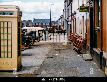 Der Fair Day in Castleisland im Jahr 1957, an dem die Bauern Vieh zum Verkauf brachten und Pferdehandel an der Tagesordnung waren, wobei die Preise angeboten/abgelehnt und schließlich abgerechnet wurden. Dieses Bild zeigt eine alte Posts and Telegraph Phone Box in ihren cremefarbenen und grünen Farben, einen Nolans Bakery Van und eine Gruppe Kühe, die auf dem Fußweg stehen. Stockfoto