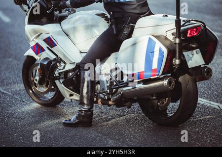 Ein Motorradpolizist in Uniform mit schwarzen Lederstiefeln steht neben einem weißen Polizeimotorrad auf einer asphaltierten Straße. Das Motorrad ist mit ausgestattet Stockfoto