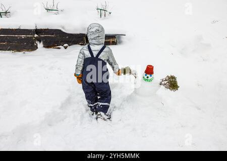 Das Kind in Winterkleidung steht neben einem kleinen Schneemann, der mit rotem Topfhut und immergrünen Zweigen verziert ist. Schweden. Stockfoto