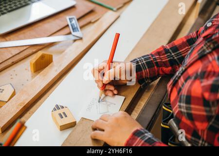 Person in einem roten karierten Hemd, die mit einem roten Bleistift Tischpläne auf Papier skizziert, umgeben von Holz Stockfoto