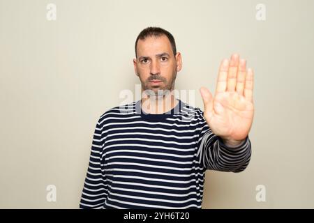 Porträt eines bärtigen hispanischen Mannes in seinen 40ern, der einen gestreiften Pullover trägt und mit der Handfläche eine Stoppgeste macht, um Stress und Probleme abzuwehren Stockfoto