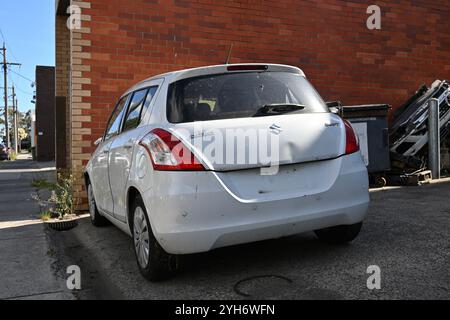 Rückansicht eines verbeulten und beschädigten weißen Suzuki Swift, der an einem sonnigen Tag neben einem Gebäude in einem Industriegebiet parkt Stockfoto