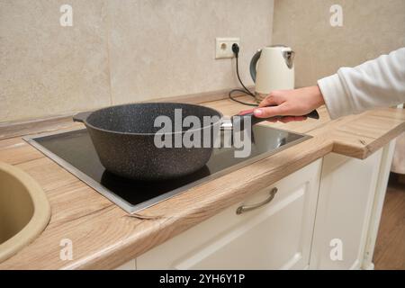 Die Hand hält die Pfanne auf einem elektrischen Herd in der modernen Küche. Inneneinrichtung mit Kochgerät und Holzarbeitsfläche. Wohnküche Konzept für Stockfoto