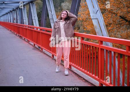 Ein einsames Weibchen steht auf einer hohen Metallbrücke, über der sich der Herbsthimmel erstreckt. Rot gekleidet posiert eine Erwachsene Frau auf einer Brücke mit kräftigen Balken, c Stockfoto