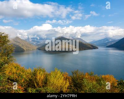 Hintergrundszene mit grünen, roten und gelben Bäumen vor dem Zhinvali-Reservoir auf dem Aragvi-Fluss an sonnigem Tag mit blauem Himmel und Wolken, Herbst. Zhinval Stockfoto