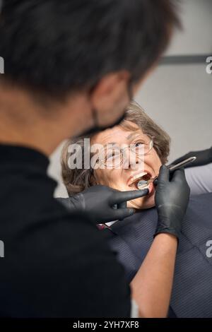 Der Zahnarzt untersucht die Zähne einer älteren Frau mit einem Zahnspiegel Stockfoto