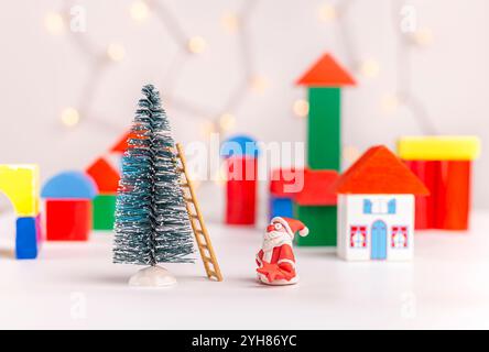 Der Weihnachtsmann aus Plastilin mit einem Stern in der Hand will einen Weihnachtsbaum auf einem verschwommenen Hintergrund eines Dorfes aus Holzwürfeln schmücken. Weihnachten Stockfoto