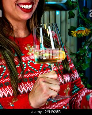 Schuss einer jungen Frau, die lächelt, während sie ein Glas mit Weißwein oder Champagner in einem roten Weihnachtspullover hält. Konzept Weihnachtsfeierlichkeiten Stockfoto