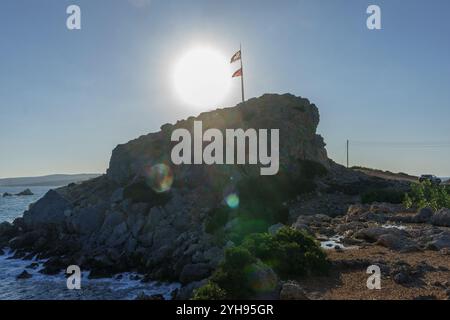 Die Sonne scheint hell über einem felsigen Felsvorsprung mit einer winkenden Flagge und überblickt die ruhigen Küstengewässer. Stockfoto