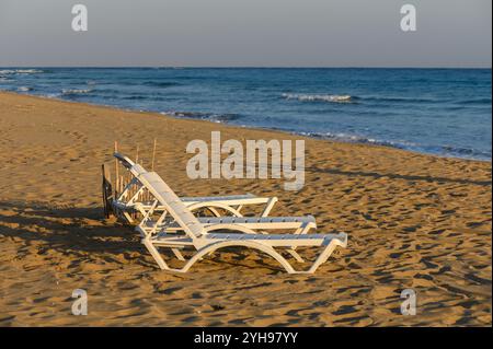 Zwei weiße Liegestühle liegen auf warmem goldenem Sand und blicken auf die sanften Wellen, während die Sonne am Horizont untergeht. Stockfoto