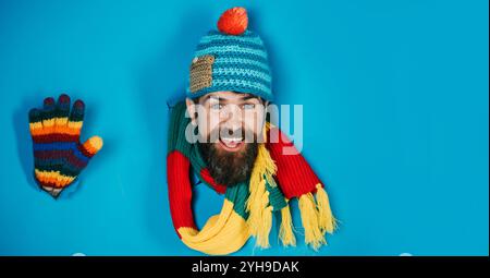 Lächelnder bärtiger Mann, der durch ein Loch in Papier schaut und die Hand winkt. Hübscher Mann in Strickmütze, farbenfrohem Schal und Handschuhen. Modische warme Kleidung Stockfoto