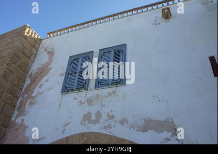 Malerische blaue Fensterläden unterstreichen die rustikale Schönheit einer verwitterten Mauer in einem historischen Küstendorf unter klarem Himmel. Stockfoto