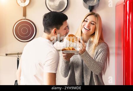 Spaß beim Frühstück. Junge Frau, die ihren Mann mit Croissant in der Küche füttert Stockfoto