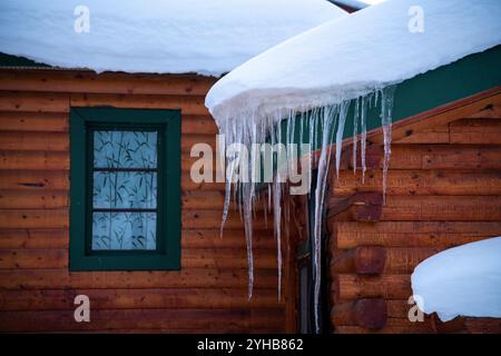 Hängende lange Eiszapfen auf der Seite einer atemberaubenden Blockhütte, Haus, Hausgebäude mit scharfen Kanten im Winter. Stockfoto