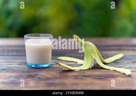 Bananensaft in einer blauen Glasschale und die Bananenschale auf einem Holztisch auf einem grünen, verschwommenen Hintergrund der Natur Stockfoto