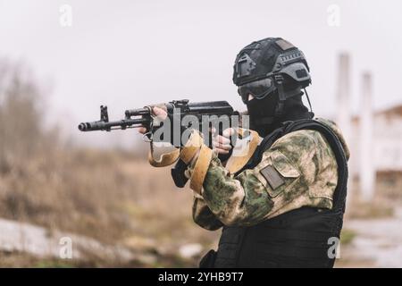 Foto eines voll ausgestatteten Soldaten in gepanzerter Weste, Helm, Gesichtsbrille und Schutz, der mit einer Waffe auf zerstörtem Stadthintergrund angreift. Stockfoto