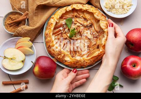 Eine Person legt vorsichtig eine frisch gebackene Apfelgalette auf einen Tisch, umgeben von geschnittenen Äpfeln, Zimtstangen und Mandelflocken. Die warme Küche Stockfoto