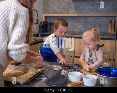 Mutter und ihre beiden Kinder kochen zusammen in der modernen Küche weihnachtskekse, indem sie Keksschneider und Pin verwenden, um fröhliche Urlaubserinnerungen zu schaffen Stockfoto