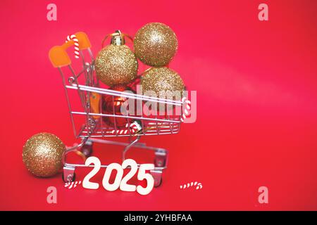 Weihnachtsdekoration Baum Spielzeug Kugeln in einem kleinen Einkaufswagen mit Girlande und Holznummern 2025 Jahre auf einem roten festlichen Hintergrund. Neujahr, Stockfoto