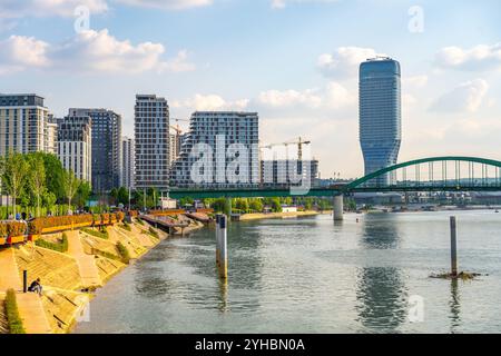 Ein lebhafter Blick auf die Sava Promenade in Belgrad mit modernen Gebäuden, üppigem Grün und einer markanten Brücke über den Fluss unter hellblauem Himmel. Stockfoto