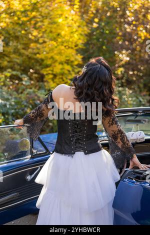 Mädchen in schwarzer Lederjacke, weißem langem geschwollenen Rock und Lederstiefeln steht neben blauem Auto in einem Herbstwald. Ihr Gesicht ist von dunklen Haaren verborgen. Stockfoto