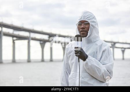 Porträt des Schwarzen Mannes, der Schutzanzug trägt und mit dem Mikrofon spricht, während er über einen Industrieunfall im Freien berichtet, der am Wasser steht und im Kopierraum ist Stockfoto