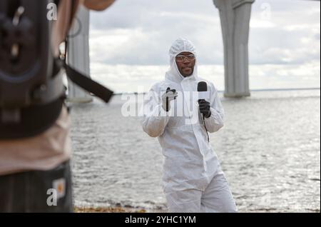 Über-Schulter-Ansicht eines Kameramanns, der Nachrichtenreportage über Industrieunfall filmt, mit Schwarzen Mann, der draußen mit Mikrofon spricht, Kopierraum Stockfoto