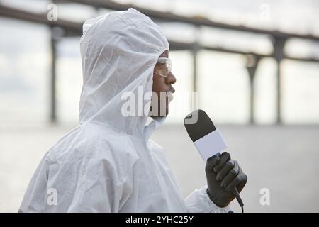 Seitliches Porträt des Schwarzen Mannes, der Schutzanzüge trägt und mit dem Mikrofon spricht, während er über Öko-Unfälle im Freien berichtet, Kopierraum Stockfoto