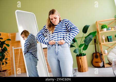 Foto einer attraktiven blonden jungen Frau gestresste Taste Anprobieren Sie neue Jeans gekleidetes gestreiftes Outfit gemütliches Tageslicht Zuhause Wohnzimmer Stockfoto