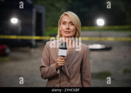 Porträt einer blonden erwachsenen Frau als Reporterin, die mit dem Mikrofon spricht, während sie den Tatort abdeckt und die Kamera und den Kopierraum ansieht Stockfoto