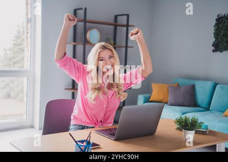 Die aufgeregte junge Frau hebt triumphierend die Arme, während sie im sonnigen Wohnzimmer ferngesteuert am Laptop arbeitet Stockfoto