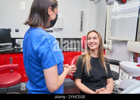 Eine Frau in einem schwarzen Hemd sitzt in einem Zahnarztstuhl, während eine Frau in einem blauen Hemd mit ihr spricht Stockfoto