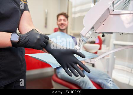 Junger lateinischer Zahnarzt in blau gekleidet, Details an Handschuhen, Hintergrund der Zahnarztkabine mit sitzendem Klienten, nicht erkennbares Modell Stockfoto