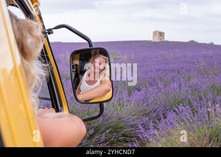 Autospiegelspiegelspiegelung einer jungen lächelnden blonden Frau, die während des Sonnenuntergangs von ihrem gelben Retro-Wohnmobil aus den Lavendelblumenfeldern aus genießt Stockfoto