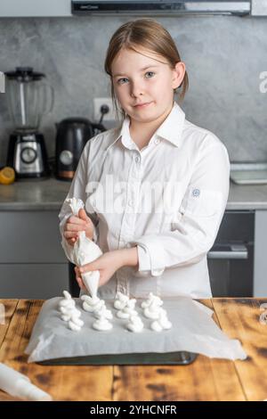 Niedliches kleines Gebäck-Mädchen in einer weißen Jacke, das einen Paspelbeutel in den Händen hält und Meringue auf Backpapier drückt. Vertikales Foto. Hohe Qualität Stockfoto