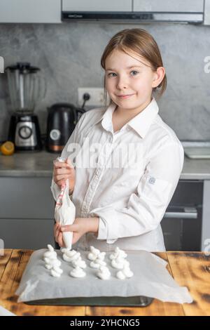 Niedliches kleines Gebäck-Mädchen in einer weißen Jacke, das einen Paspelbeutel in den Händen hält und Meringue auf Backpapier drückt. Vertikales Foto. Hohe Qualität Stockfoto