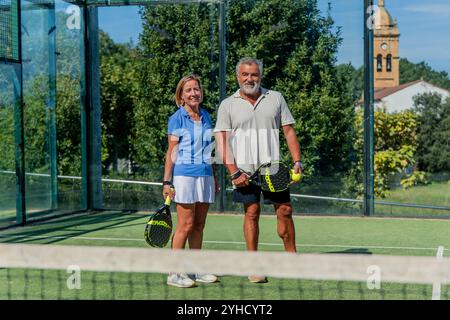 Ältere Padel-Spieler stehen auf dem Spielfeld und blicken entspannt in die Kamera, während sie ihre Paddel halten Stockfoto